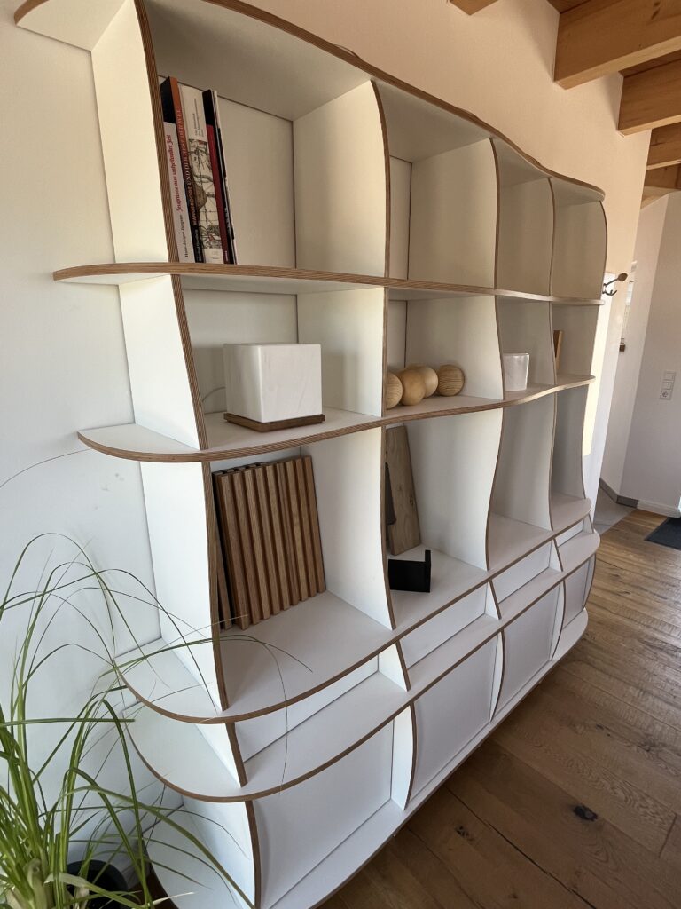 Designer-Regal aus weißem Multiplex mit geschwungenen Seitenwänden und offenen Fächern, dekoriert mit Holzobjekten, Büchern und einer Zimmerpflanze im modernen Wohnraum